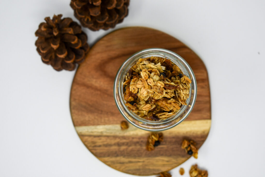 picture from above of granola in jar on top of wooden plate with pinecones next to it