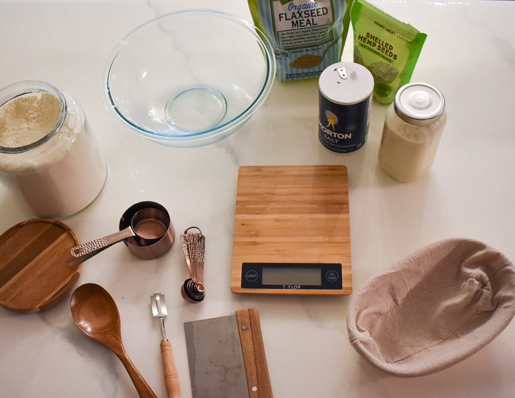 Picture of a glass bowl, flour, kitchen scale, banneton, measuring cups, measuring spoons, wooden spoon, salt, hemp seeds, flax seed, and sourdough starter on a marble countertop.