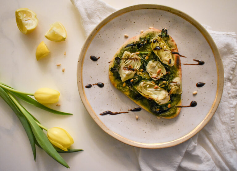 artichoke and pesto pizza on a gray plate on top of a white towel, surrounded by yellow tulips and lemon wedges